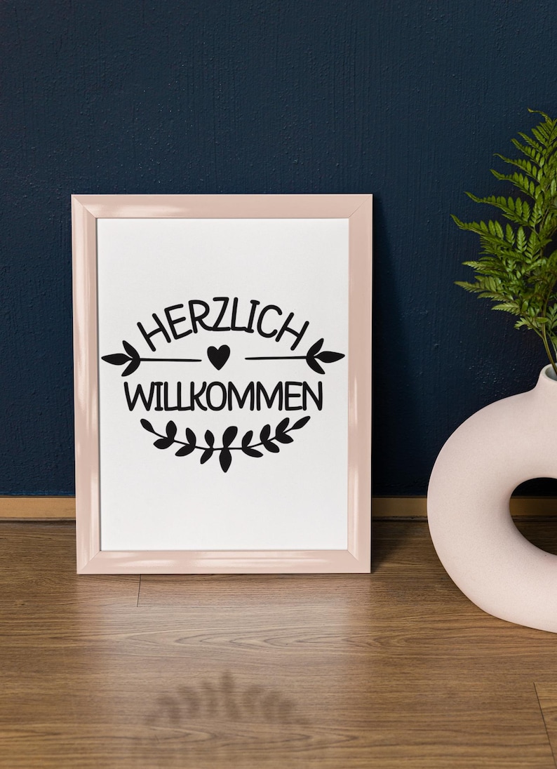 Op de afbeelding: Een roze ingelijste print met de tekst "HERZLICH WILLKOMMEN" in zwarte letters met een klein hartje en een simpel pijlontwerp. De print is omringd door een krans van bladeren.
