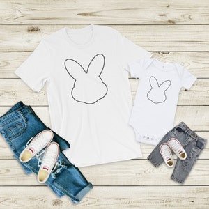 Peut inclure: Deux t-shirts blancs avec un contour simple de lapin sur le devant. Le plus grand t-shirt a un motif de lapin plus grand. Le plus petit t-shirt est une grenouill&egrave;re. Les deux t-shirts sont dispos&eacute;s sur une surface en bois avec un jean et des baskets.