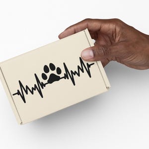 May include: A beige cardboard box featuring a black paw print design integrated into a heartbeat line. The box is held by a hand against a white background. The design suggests a pet-related theme.