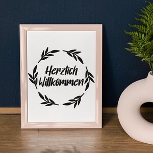 Op de afbeelding: Een ingelijst afdruk met een roze lijst en een witte achtergrond. De afdruk toont een zwarte krans van bladeren met de tekst "Herzlich Willkommen" in het midden.