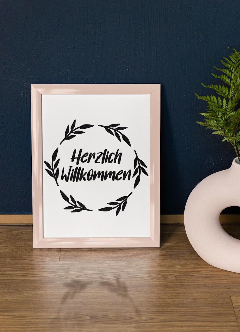 Op de afbeelding: Een ingelijst afdruk met een roze lijst en een witte achtergrond. De afdruk toont een zwarte krans van bladeren met de tekst "Herzlich Willkommen" in het midden.