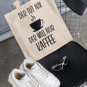 以下が含まれることがあります： 「DER TUT NIX DER WILL NUR KAFFEE」の文字とコーヒーカップのグラフィックが入ったベージュのトートバッグ。また、白いスニーカー、濃いグレーのビーニー、シルバーフレームのサングラスが椅子の上に置かれています。