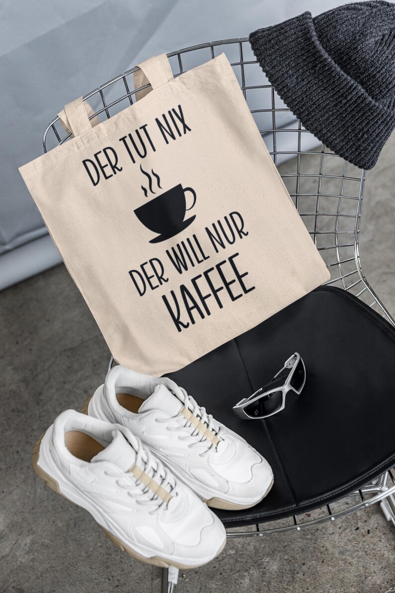 以下が含まれることがあります： 「DER TUT NIX DER WILL NUR KAFFEE」の文字とコーヒーカップのグラフィックが入ったベージュのトートバッグ。また、白いスニーカー、濃いグレーのビーニー、シルバーフレームのサングラスが椅子の上に置かれています。
