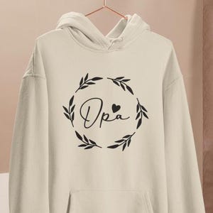 May include: A white hoodie with a black design of a wreath with the word "Opa" and a small heart in the centre.