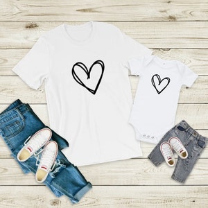 May include: Two white t-shirts with a black hand-drawn heart design. The larger shirt has a larger heart design. The smaller shirt is a baby onesie. The shirts are laid out on a light wood background with blue jeans and white sneakers.