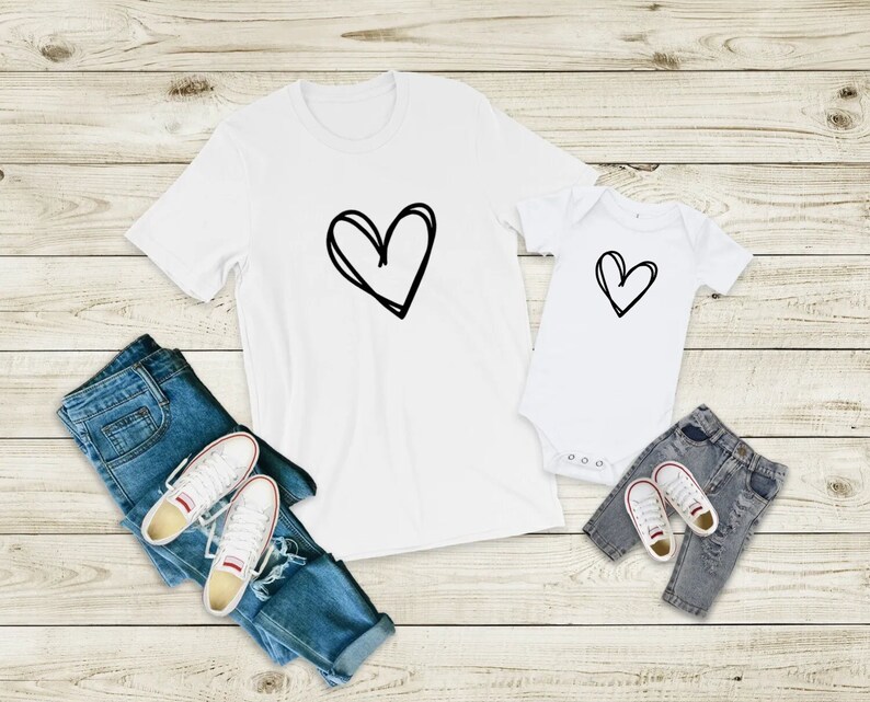 May include: Two white t-shirts with a black hand-drawn heart design. The larger shirt has a larger heart design. The smaller shirt is a baby onesie. The shirts are laid out on a light wood background with blue jeans and white sneakers.