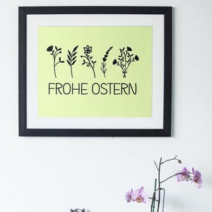 May include: A framed print with a light green background and black silhouettes of flowers. The text "FROHE OSTERN" is printed in black below the flowers.