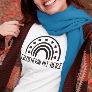 May include: White t-shirt with a black rainbow design and the text "ERZIEHERIN MIT HERZ". The person is wearing a blue scarf and a brown cardigan.
