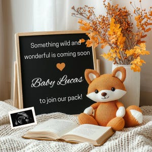 Pode incluir: Uma lousa preta com o texto "Something wild and wonderful is coming soon. Baby Lucas to join our pack!" Um pequeno brinquedo de pelúcia de raposa laranja está sentado na frente da lousa. Uma imagem de ultrassom e um livro aberto também estão na imagem.