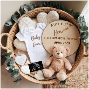 Pode incluir: Uma placa de madeira com o texto "A little bit of Heaven fell from above arriving April 2025" em uma cesta de vime com um macacão branco que diz "Baby Emma", um urso de pelúcia e uma imagem de ultrassom.