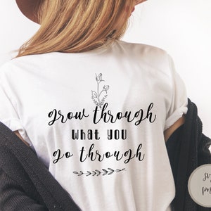 Op de afbeelding: Wit T-shirt met de tekst "grow through what you go through" in zwarte cursieve letters. Boven de tekst staat een klein bloemetjesmotief. Het shirt wordt door iemand gedragen.