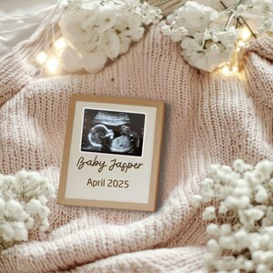 Pode incluir: Uma moldura de madeira com fundo branco e uma imagem de ultrassom em preto e branco de um bebê. O texto "Baby Jasper April 2025" está escrito em letras cursivas marrons.