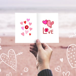 K&ouml;nnte beinhalten: Eine wei&szlig;e Karte mit rosa und roten Herzen und einem rosa Vogel mit dem Wort "LOVE" in rot. Die Karte wird von einer Hand vor einem verschwommenen Hintergrund eines Strandes mit wei&szlig;en Herzen gehalten, die in den Sand gezeichnet sind.