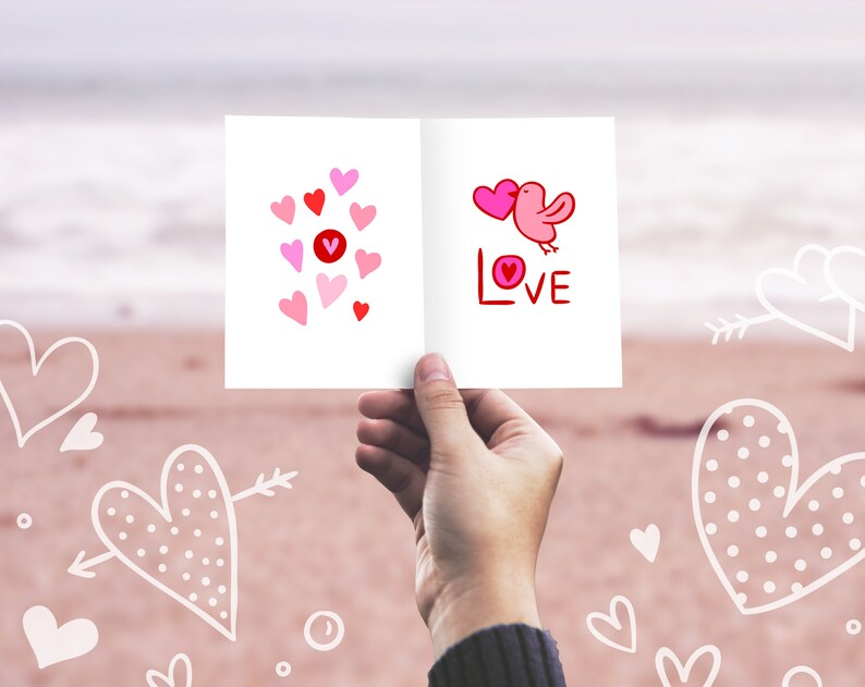 K&ouml;nnte beinhalten: Eine wei&szlig;e Karte mit rosa und roten Herzen und einem rosa Vogel mit dem Wort "LOVE" in rot. Die Karte wird von einer Hand vor einem verschwommenen Hintergrund eines Strandes mit wei&szlig;en Herzen gehalten, die in den Sand gezeichnet sind.