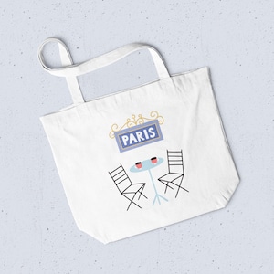 May include: White tote bag with a Parisian cafe design. It features a blue sign reading "PARIS", a small table, two chairs, and two coffee cups. The bag has two handles for carrying.
