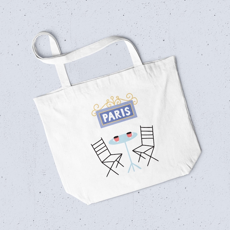 May include: White tote bag with a Parisian cafe design. It features a blue sign reading "PARIS", a small table, two chairs, and two coffee cups. The bag has two handles for carrying.