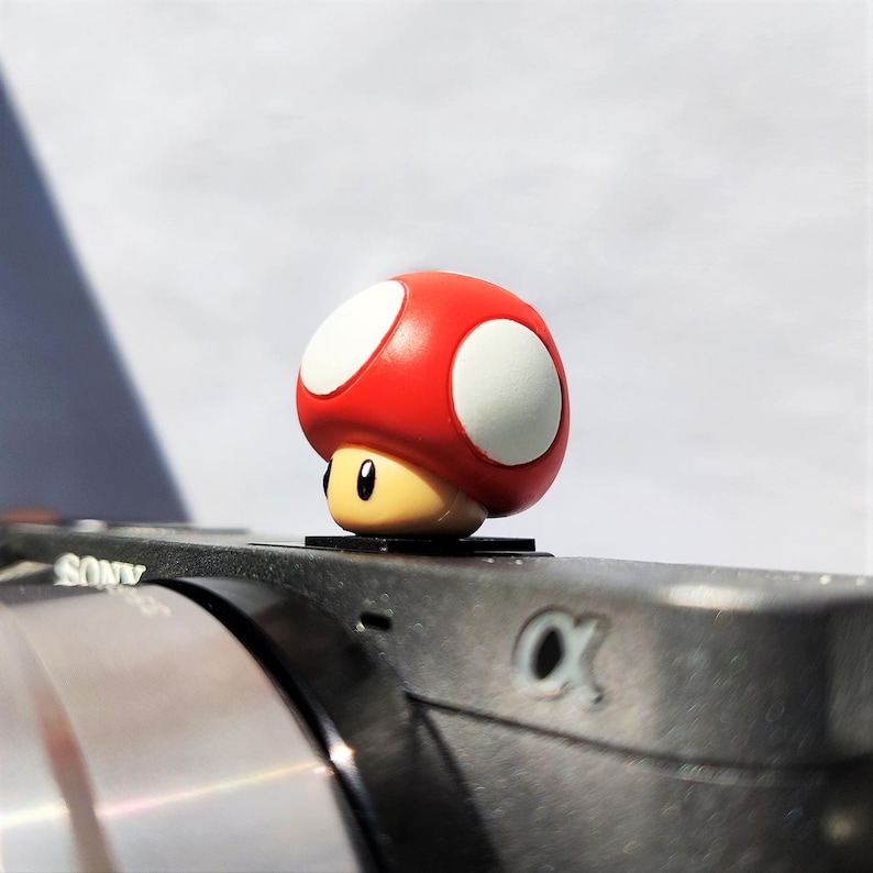 May include: A red and white mushroom-shaped hot shoe cover featuring a smooth, rounded cap with white spots and a tan base with black eyes, mounted onto the top of a black camera body with the Sony brand name and alpha symbol visible.