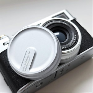 May include: A silver X100VI camera lens cap made of aluminum alloy sits next to a camera lens. The lens is black with silver accents and has the text "EBC f=23mm 1:2". The camera body is silver and black.