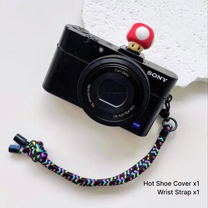 May include: A black Sony digital camera with a Carl Zeiss Vario-Sonnar lens, featuring a red and white mushroom hot shoe cover and a braided wrist strap with a multi-coloured chevron pattern in black, blue, pink, and yellow, plus text: Hot Shoe Cover x1, Wrist Strap x1.