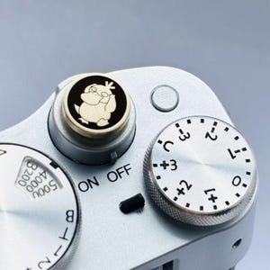 May include: Close-up of a silver camera body with various dials and controls. A black and gold dial features a cartoon character. The camera has settings for ISO, shutter speed, and exposure compensation. The text "ON OFF" is visible.