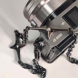 May include: A black chain camera strap with a silver camera attached. The camera strap has two star-shaped charms with rhinestones. The camera is a Sony Alpha 6000.