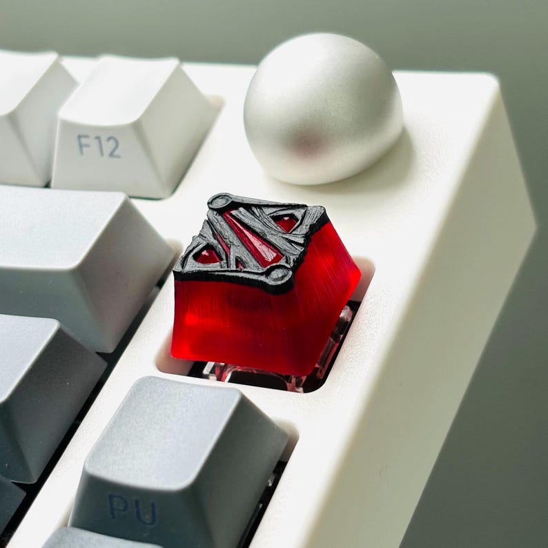May include: A red and black mechanical keyboard keycap with a stylized logo. The keycap is on a white keyboard.