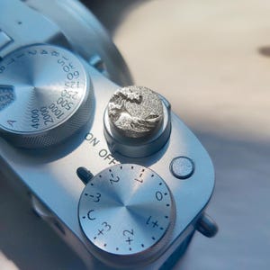 May include: A silver-toned camera shutter button cover featuring a raised, textured relief of a crashing wave design. The circular accessory is mounted on the top of a silver camera body, positioned next to a circular dial with numerical markings.