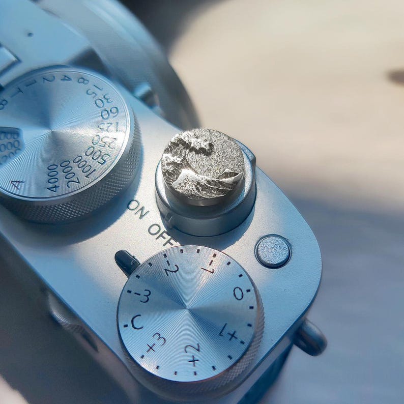 May include: A silver-toned camera shutter button cover featuring a raised, textured relief of a crashing wave design. The circular accessory is mounted on the top of a silver camera body, positioned next to a circular dial with numerical markings.