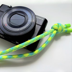 May include: Black Sony digital camera with a Carl Zeiss lens. A colorful camera strap in a pattern of yellow and blue-green is knotted and attached to the camera. The strap ends are bright green.