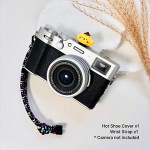 May include: A silver and black digital camera featuring a yellow cartoon character hot shoe cover and a multicolored braided wrist strap. The text on the image reads: Hot Shoe Cover x1, Wrist Strap x1, * Camera not included.