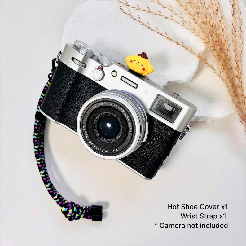 May include: A silver and black digital camera featuring a yellow cartoon character hot shoe cover and a multicolored braided wrist strap. The text on the image reads: Hot Shoe Cover x1, Wrist Strap x1, * Camera not included.