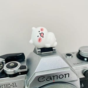 May include: A white figurine of a cartoon character with pink cheeks and a question mark on its belly rests on a silver Canon camera. The camera has black accents and the Canon logo is visible.