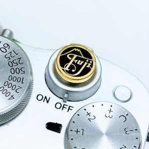 May include: A silver camera with a black and gold Fuji logo on the top dial. The camera has a silver dial with the words "ON OFF" and a silver dial with numbers and symbols.