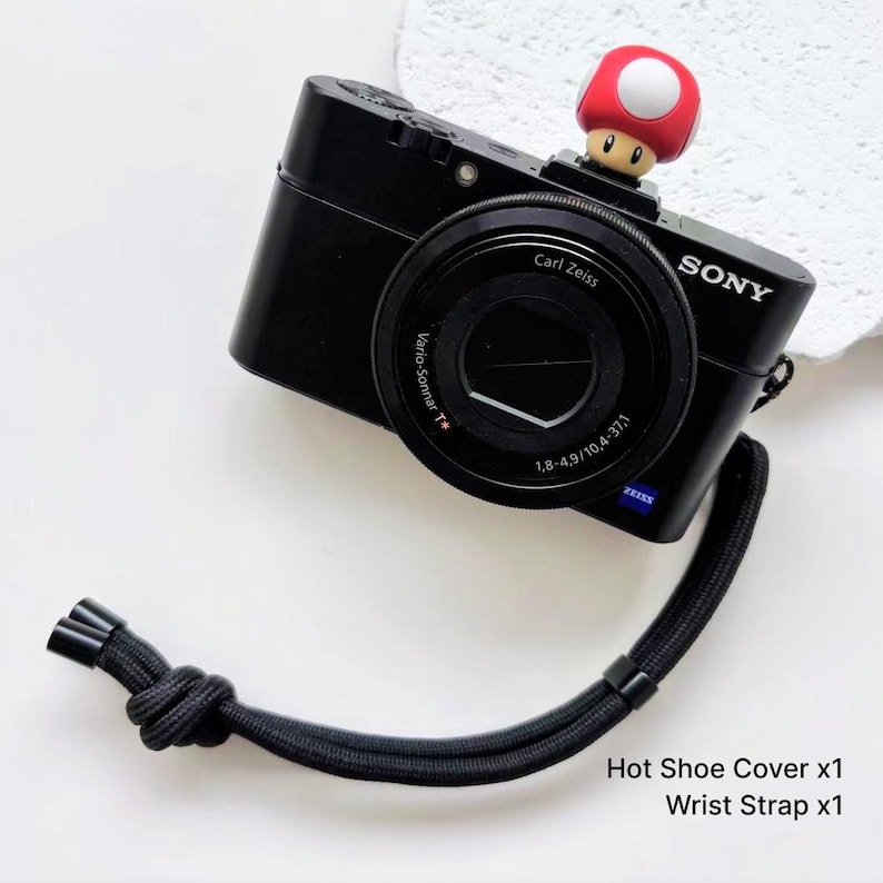 May include: A black Sony digital camera featuring a Carl Zeiss Vario-Sonnar T* lens, equipped with a red and white mushroom-shaped hot shoe cover and a black woven wrist strap. The text Hot Shoe Cover x1 and Wrist Strap x1 is visible.