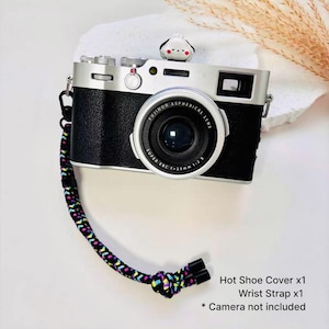 May include: A black and silver digital camera with a Fujinon lens, featuring a small white and black animal-shaped hot shoe cover on top and a braided wrist strap with a multi-colored geometric pattern in black, yellow, pink, and blue.