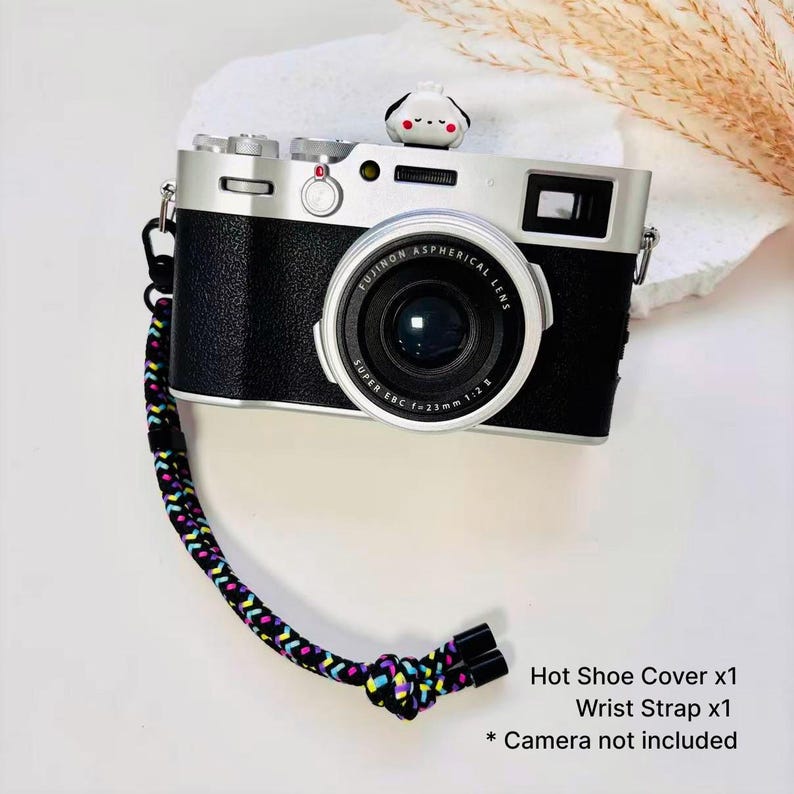 May include: A black and silver digital camera with a Fujinon lens, featuring a small white and black animal-shaped hot shoe cover on top and a braided wrist strap with a multi-colored geometric pattern in black, yellow, pink, and blue.