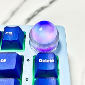 Op de afbeelding: Een blauw en wit toetsenbord met een ronde, veelkleurige, gloeiende knop op de "Delete"-toets.