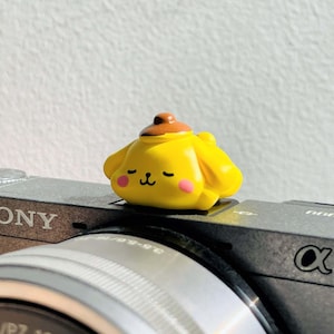 May include: A yellow figurine of a cartoon character with a brown hat and pink cheeks, resting on a camera. The figurine has closed eyes and a smiling expression. The camera is gray and has the word "SONY" printed on it.