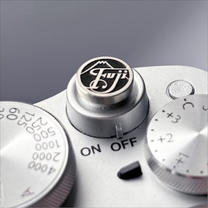 May include: Close-up of a silver camera with a black "Fujifilm" logo on the top dial. The camera has a silver dial with "ON OFF" printed on it and a silver dial with numbers for exposure compensation.