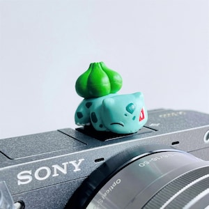 Może przedstawiać: Mała, jasnoniebieska figurka Bulbasaur z zielonymi strukturami w kształcie cebuli na plecach, spoczywająca na szarej kamerze Sony. Figurka ma ciemnozielone plamki i czerwone akcenty. Widoczny jest obiektyw aparatu.