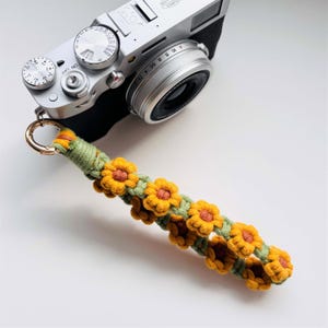 Puede incluir: Una correa de cámara de macramé con un patrón repetido de girasoles amarillos y naranjas. La correa tiene una base verde y un cierre de metal dorado. Una cámara plateada está en el fondo.