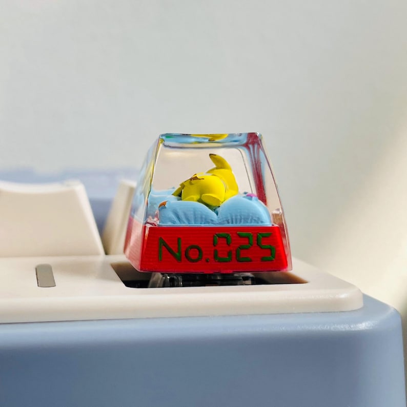 May include: A custom keycap featuring a yellow cartoon character resting on blue pillows inside a clear, rectangular enclosure. The keycap has a red base with the text "No. 025" in green.