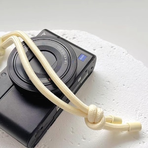 May include: A black digital camera with a cream-coloured rope strap. The camera lens has a silver ring and the strap is knotted. The camera is resting on a white surface.