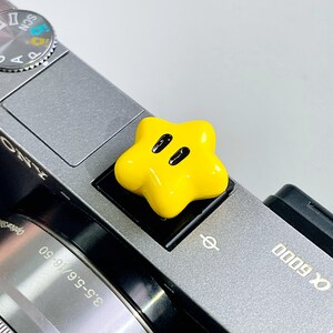 May include: A bright yellow, star-shaped camera hot shoe cover featuring two black oval eyes, attached to the top of a grey Sony Alpha 6000 camera body. The accessory has a smooth, glossy finish and is positioned over the camera's flash mount.