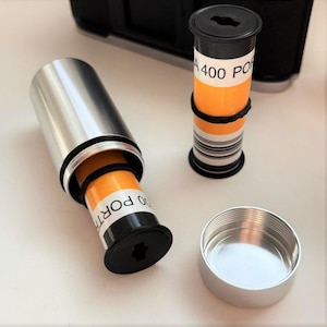 May include: A silver film canister with a black cap, next to a roll of film with the text "A400 PORT". The film is orange and white. The canister is open, revealing the film inside. A second film roll is standing upright.