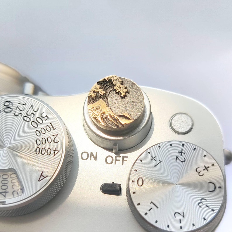 May include: A circular metal camera shutter release button featuring a gold-toned relief of a crashing wave. The button is mounted on the top plate of a silver camera, positioned between a shutter speed dial and an exposure compensation dial.