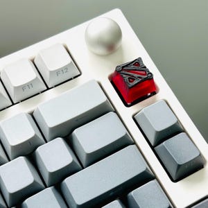 May include: A white mechanical keyboard with a red and black keycap featuring a stylized logo. The keycap is in the top right corner of the keyboard.