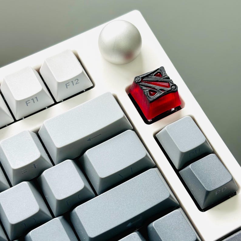 May include: A white mechanical keyboard with a red and black keycap featuring a stylized logo. The keycap is in the top right corner of the keyboard.