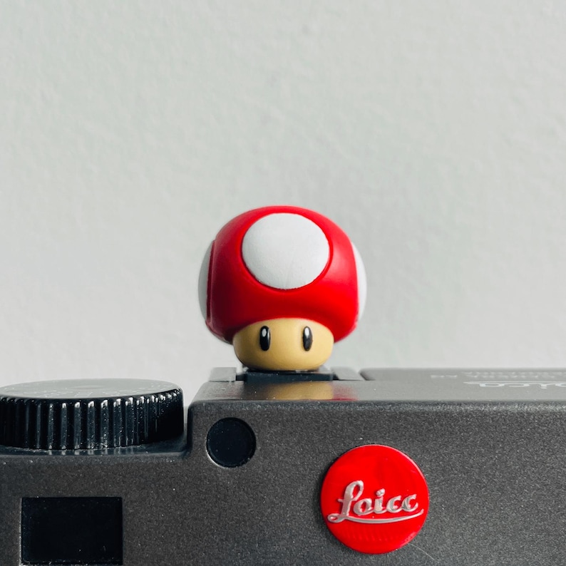 May include: A small red and white mushroom-shaped figure with a tan face and black eyes, positioned on top of a dark grey camera body. The camera features a circular red logo with the white text Leica printed in a cursive script on the front.