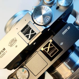 May include: A silver and black digital camera with a silver lens and a black lens cap. The camera has a black strap and a black and silver flash. The camera has a black and silver dial with the text "FUJIFILM" and "X-G000" on it.